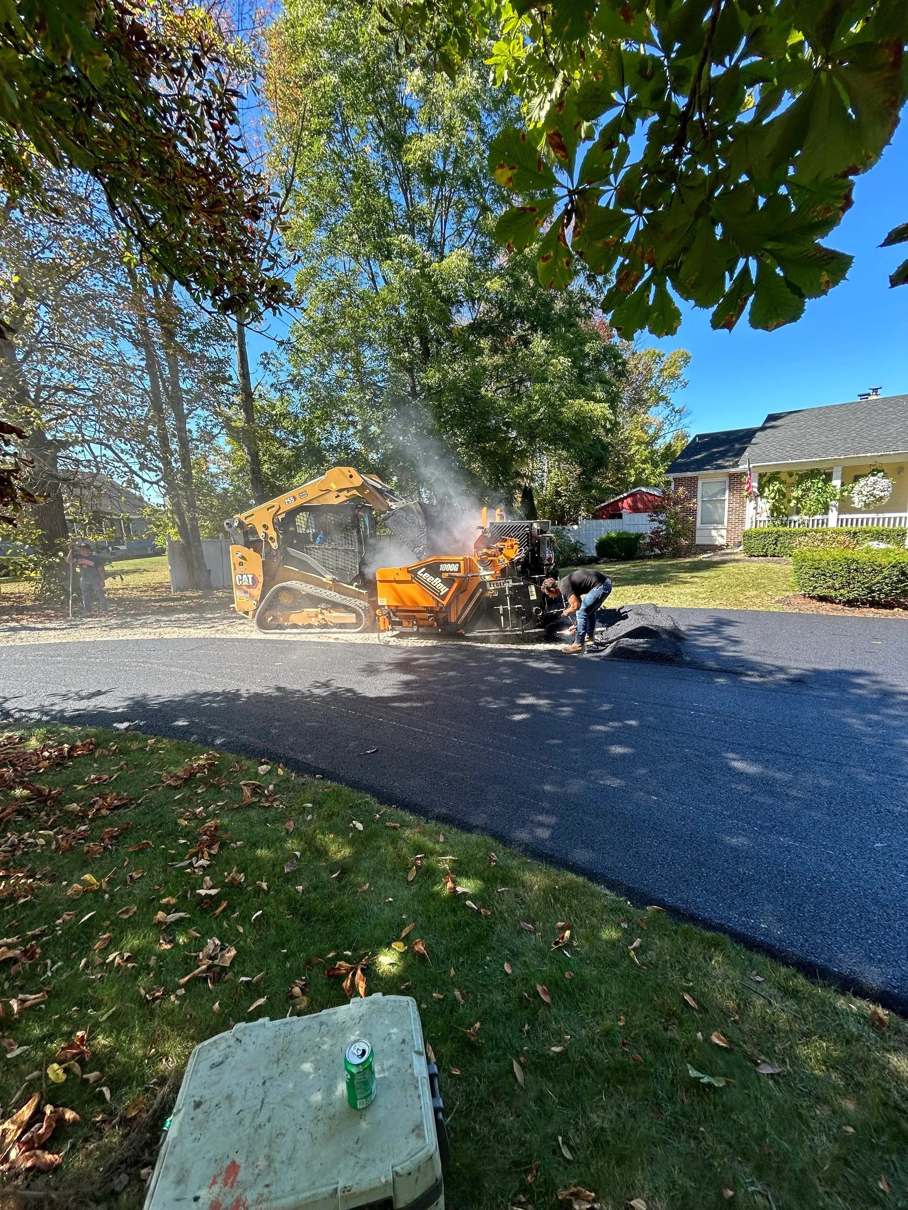 Yellow asphalt paver applying fresh asphalt to residential driveway with compaction equipment