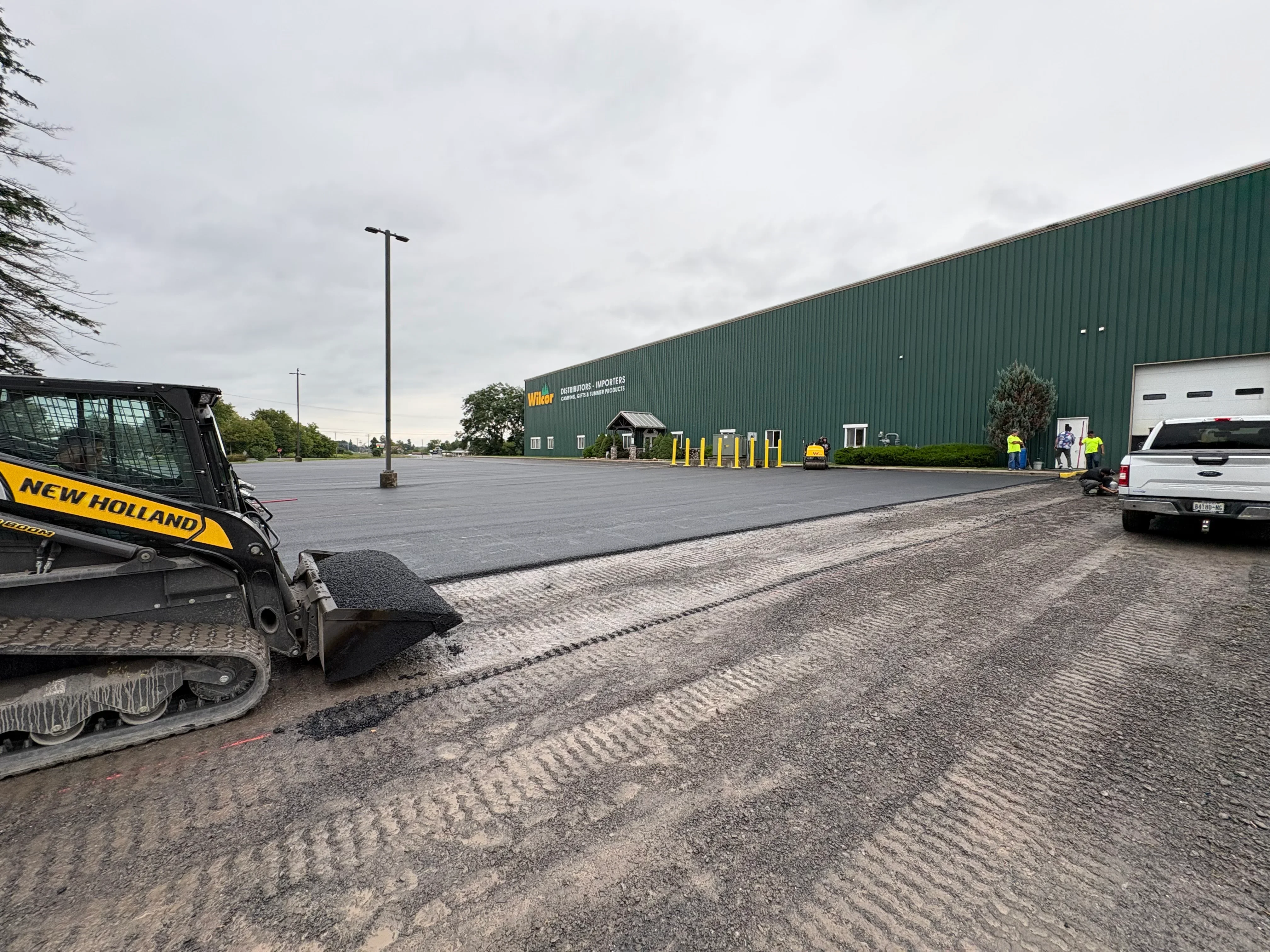 Fresh asphalt parking lot with compaction marks, yellow safety bollards, and industrial building