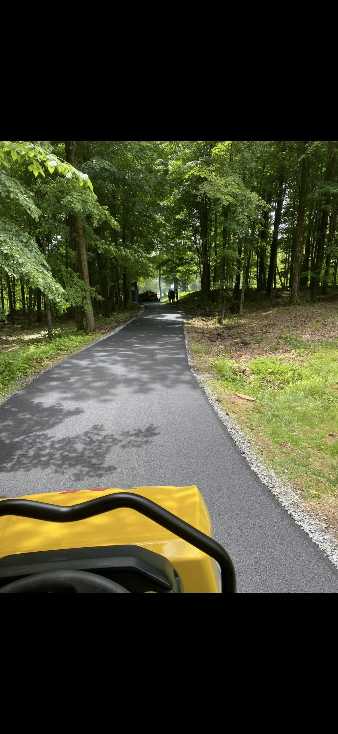 Newly paved asphalt driveway with gravel edges winding through wooded area with mature trees