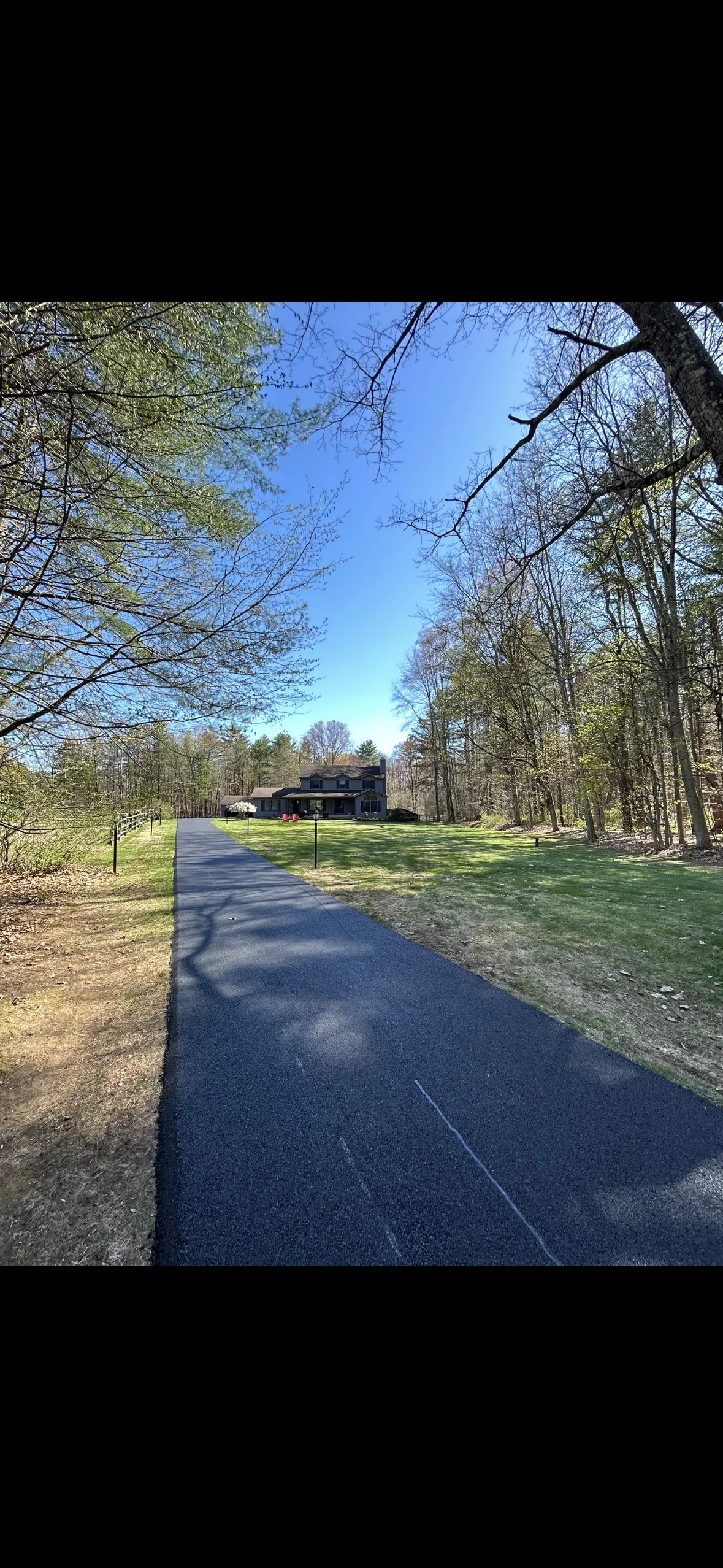 Dark asphalt driveway through wooded property with mature trees and green lawn