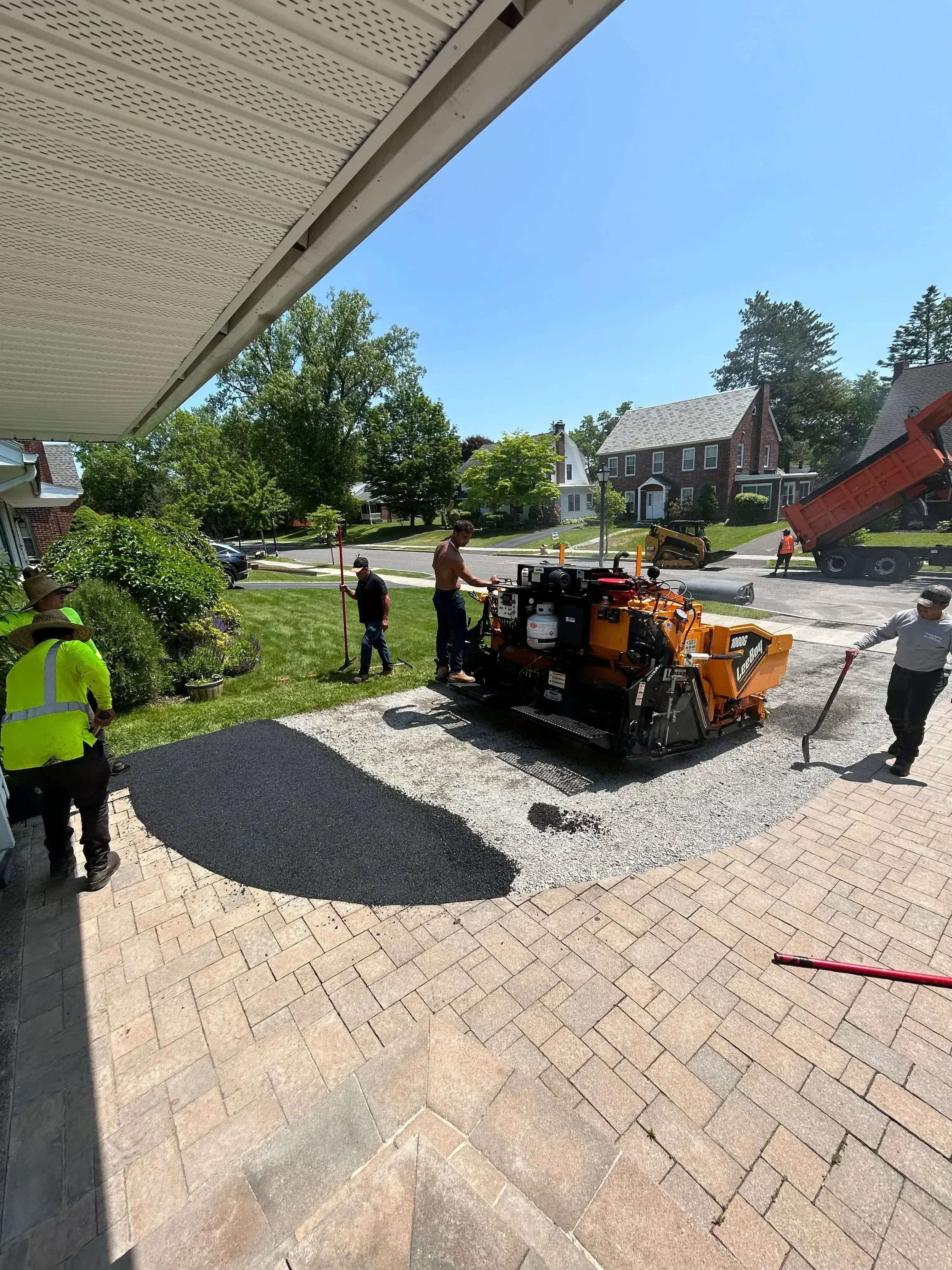 Asphalt paving equipment laying fresh black asphalt on residential driveway next to brick pavers
