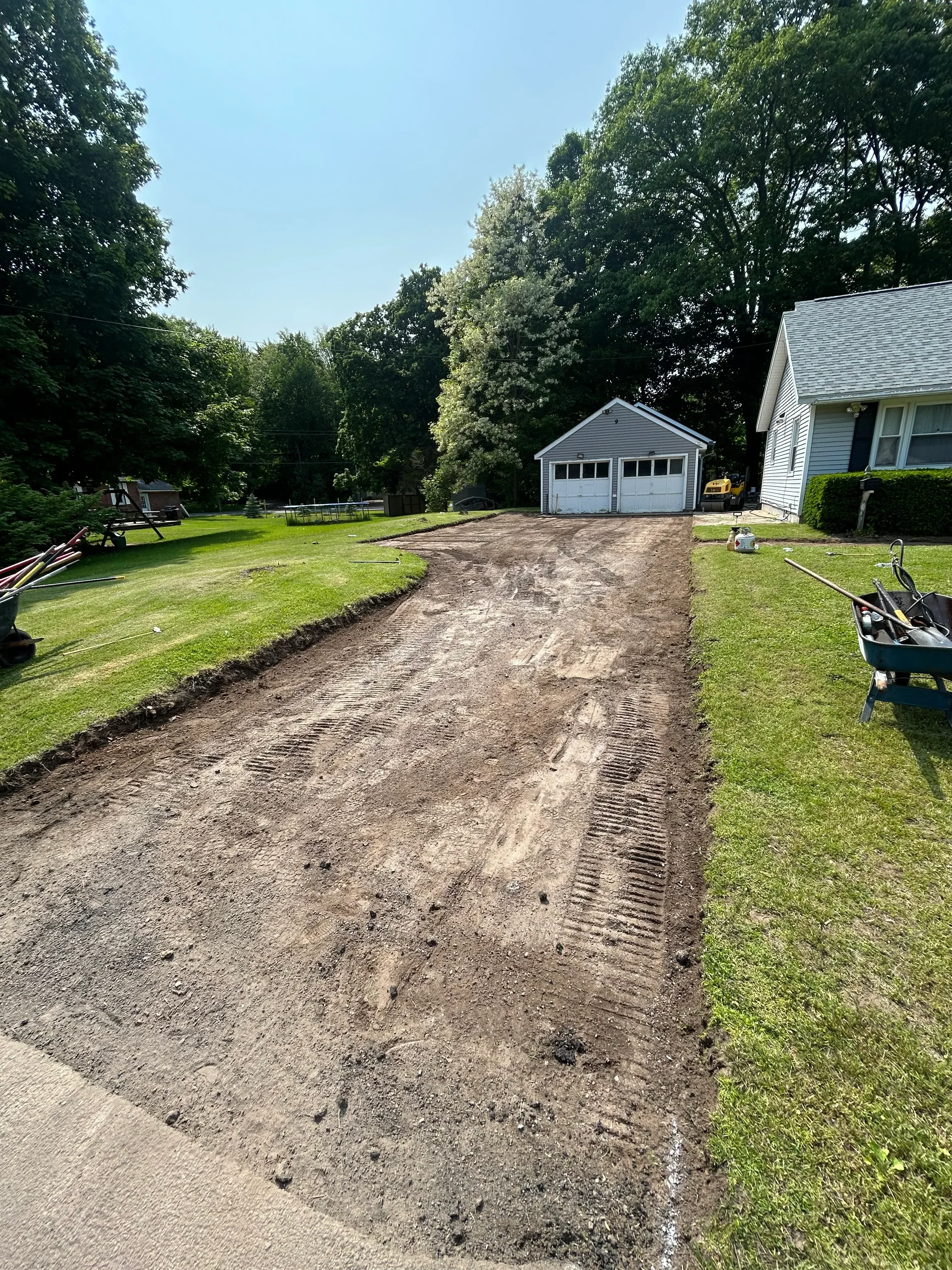 Excavated and graded driveway subbase with tractor marks, bordered by green lawn and mature trees