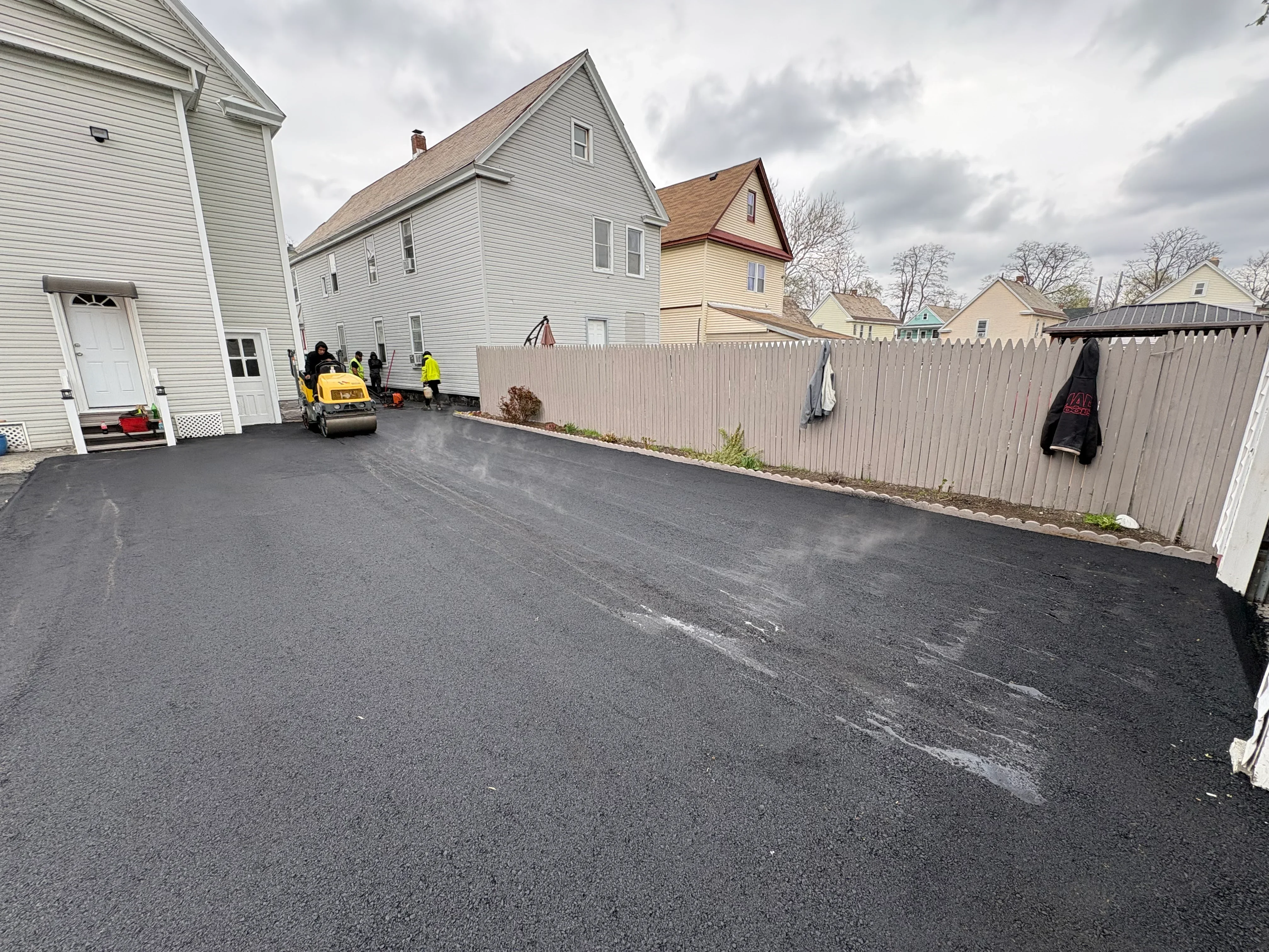 Fresh asphalt driveway installation with compaction equipment and crew on residential property