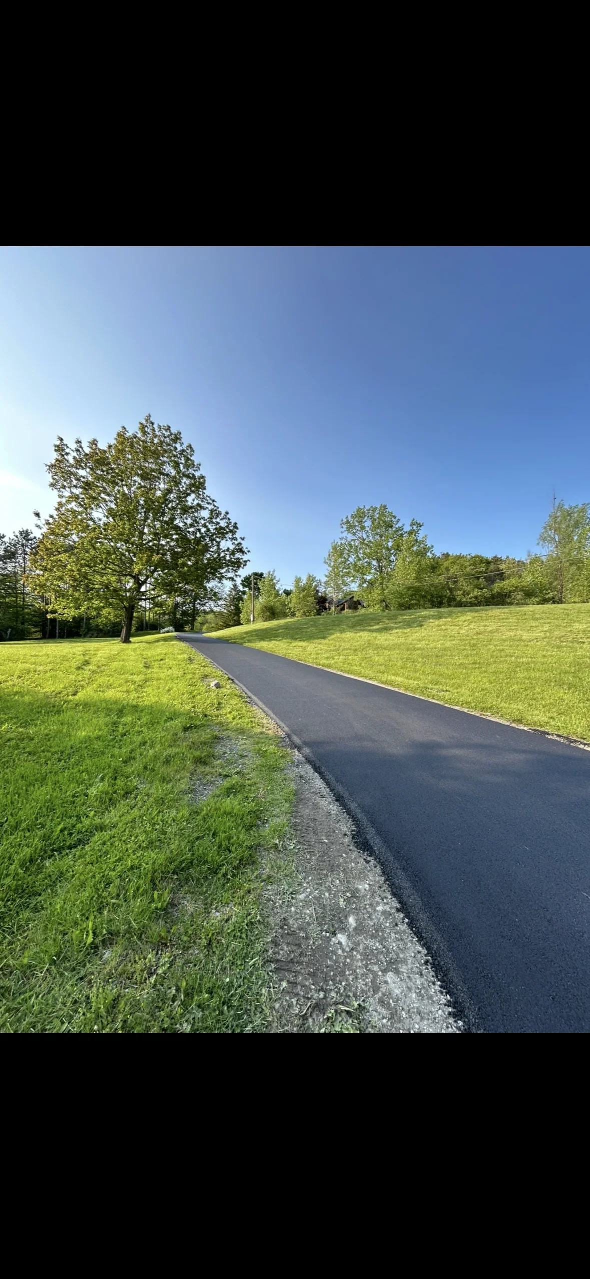 New black asphalt driveway with gravel base on sloped residential property with mature trees