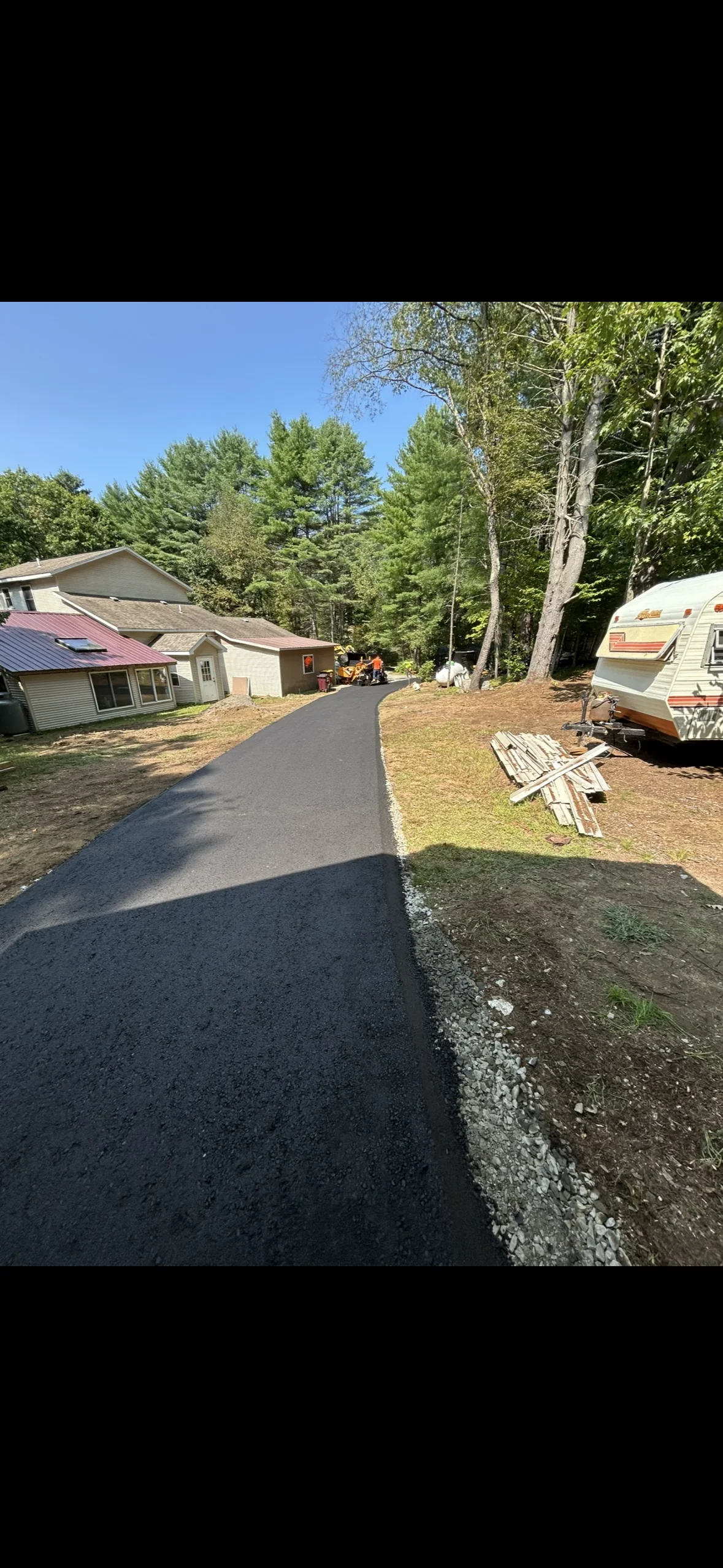 Fresh asphalt driveway with white edge line, residential home and trailer visible in wooded setting